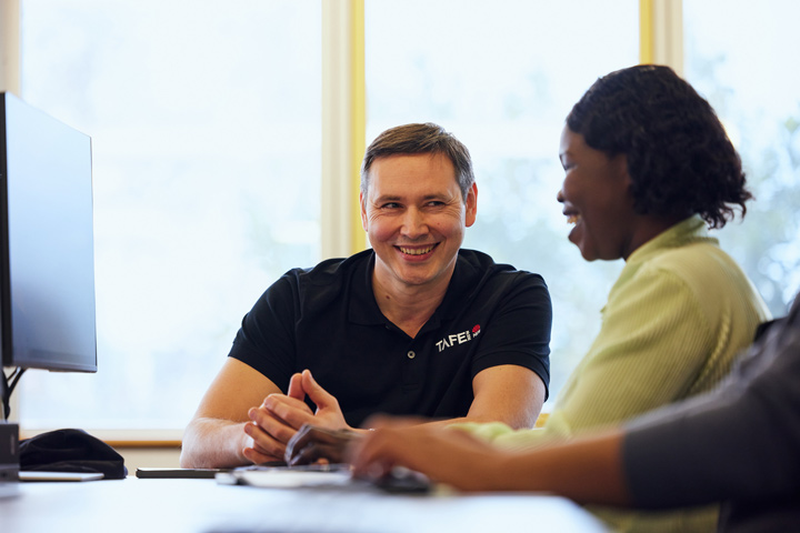 Two individuals in a library study area discussing learning support, with one wearing a black TAFE NSW branded shirt and the other in a light green sweater.