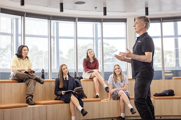 Teacher speaks to a group of school students seated on curved wooden tiered benches in the TAFE NSW amphitheatre space, with large windows providing natural light and personal belongings placed beside the students.