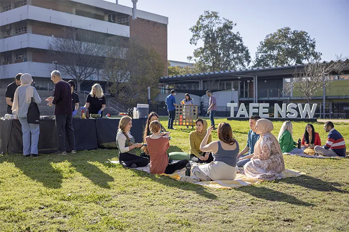 People sit on picnic blankets and gather around serving tables on a grassy area during a TAFE NSW BBQ event, with campus buildings and a large TAFE NSW sign visible in the background under bright afternoon sunlight.