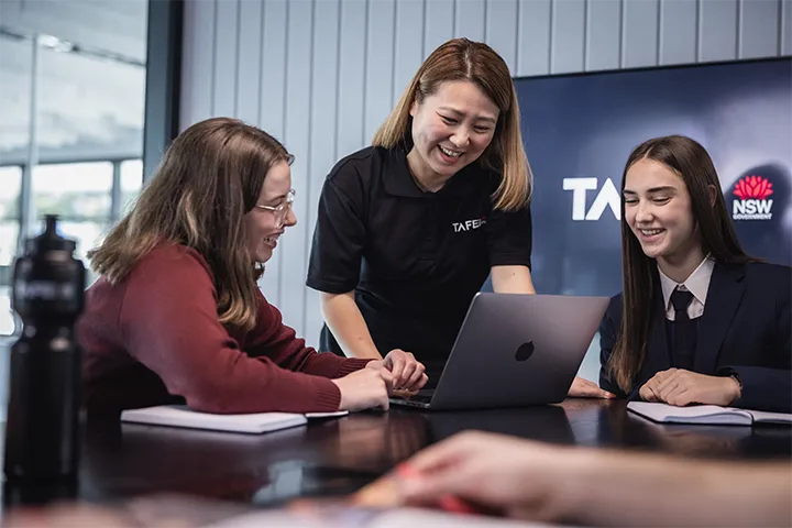 Advisor stands at a table guiding two students during a meeting, with a laptop open in front of them, notebooks on the table, and a TAFE NSW screen displayed in the background of the modern meeting room.