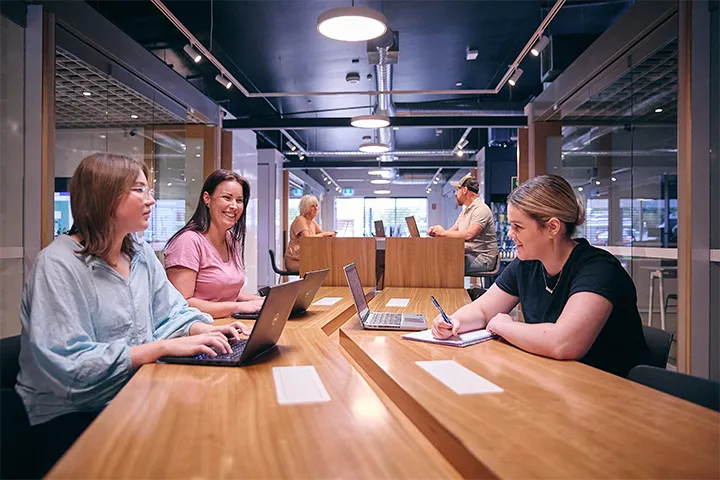 Three people participate in a boardroom meeting at TAFE NSW, seated at modern wooden tables with laptops and notebooks, engaging in collaborative discussions in a well-lit room with glass partitions.