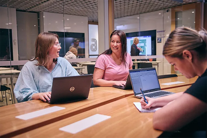 alt=”Three female students work in a glass classroom on laptops.”