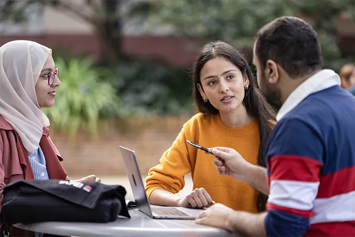 Three students sitting outdoors around a table, discussing and collaborating with a laptop and notes.