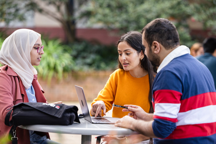 Three students sit around a small outdoor table working together on a laptop, with one person pointing to the screen while others take notes and discuss ideas, backpacks and study materials placed on the table in a leafy campus setting.