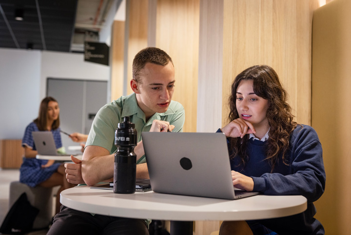 Two students collaborating at a round table in a study pod, working on a laptop with a TAFE-branded water bottle on the table, in a modern indoor study area with wooden wall paneling.