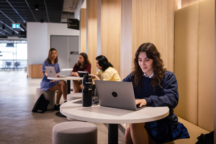 Students using study pods in a modern indoor learning space, each working on laptops at round tables, with wooden wall paneling and a TAFE-branded water bottle visible.