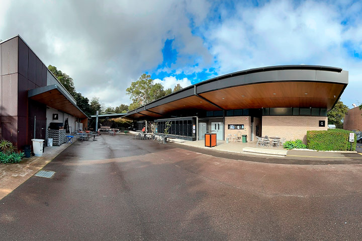 Exterior of a modern building with a curved roof, large windows, and an outdoor seating area with tables and chairs, likely part of the Design Centre Enmore. Trees are in the background and the ground appears wet under a partly cloudy sky.