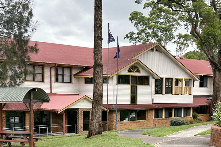 Exterior of the TAFE NSW Kempsey campus featuring a two‑storey heritage‑style building with cream weatherboard walls, red metal roofs, brick lower level, and Australian flags raised on flagpoles at the front, surrounded by mature trees, lawns, and paved walkways under an overcast sky.