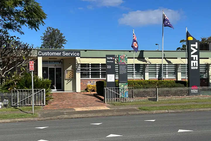 Exterior view of the TAFE NSW Wauchope campus showing a single‑storey building with green roofing, brick and rendered walls, large front windows, and three flagpoles displaying flags at the entrance, set behind a low fence with landscaped gardens and a footpath along the street.