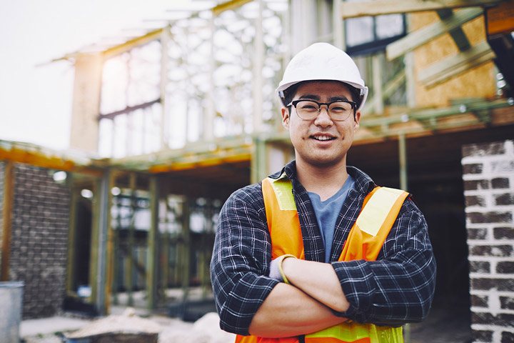 A man stands in front of a construction site, dressed in a hi-vis vest and hard hat. He wears black rimmed glasses, has his arms crossed and is smiling toward the camera.