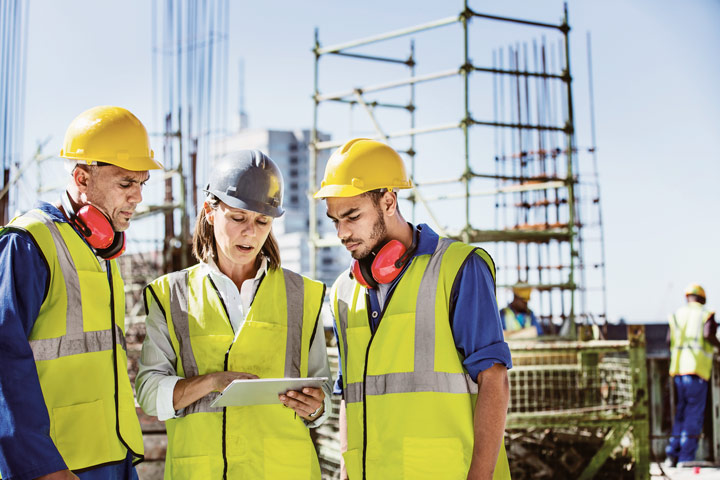 Three individuals in high-visibility vests and hard hats discussing a project at a construction site with scaffolding and workers in the background.