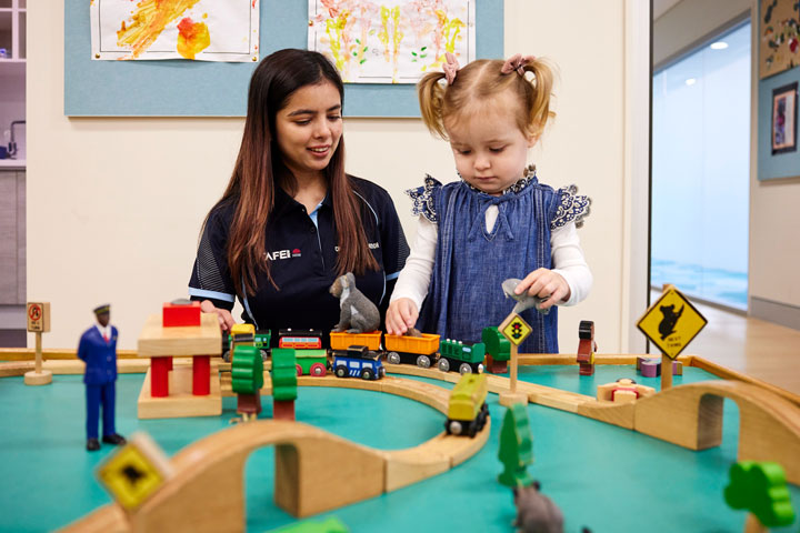 A young woman with long brown hair wears a TAFE NSW polo shirt and sits with a toddler in a childcare facility. They are sitting in front of a train track and are engaged in playing trains together.