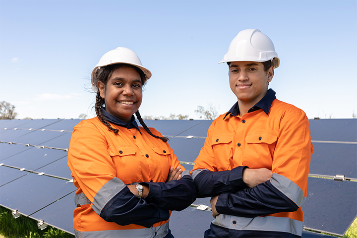 A man and a woman standing in high-vis uniforms and hard-hats are smiling at the camera. There are solar panels behind them.