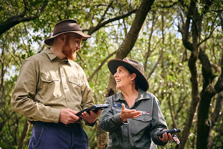 A man and woman wearing outdoor work attire and hats are smiling and having a conversation in a forested area. The man holds a tablet, while the woman gestures expressively, discussing something intently.
