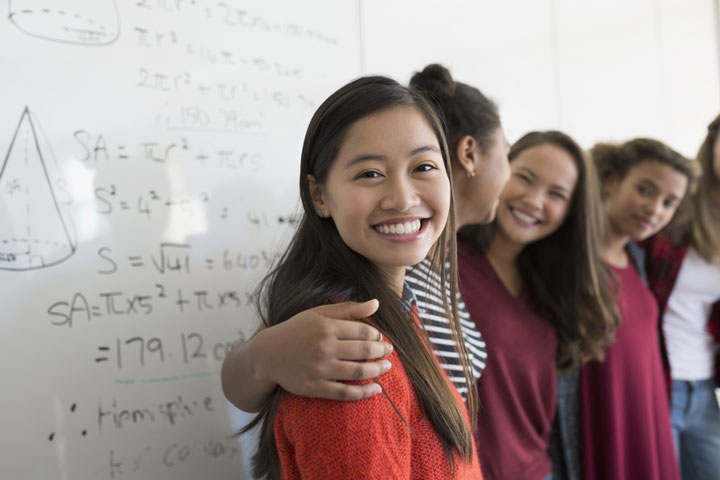 A group of women stand together side by side in front of a whiteboard. The camera is focused on one smiling young women facing the camera, with the other women blurred in the foreground. 