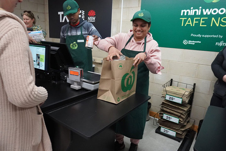 TAFE NSW students pack groceries into a paper bag at a supermarket checkout.