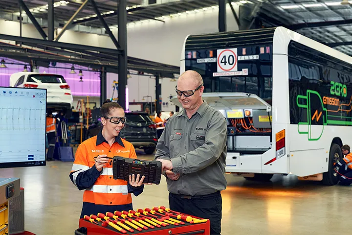 Automotive teacher guiding a female student in a workshop, both engaged in collaborative work on an iPad.