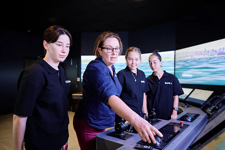 TAFE NSW maritime teacher demonstrates navigation controls to students inside the Newcastle maritime simulator, with curved floor‑to‑ceiling screens displaying an ocean and coastline scene and ship control panels positioned in front of the group for hands‑on maritime training.