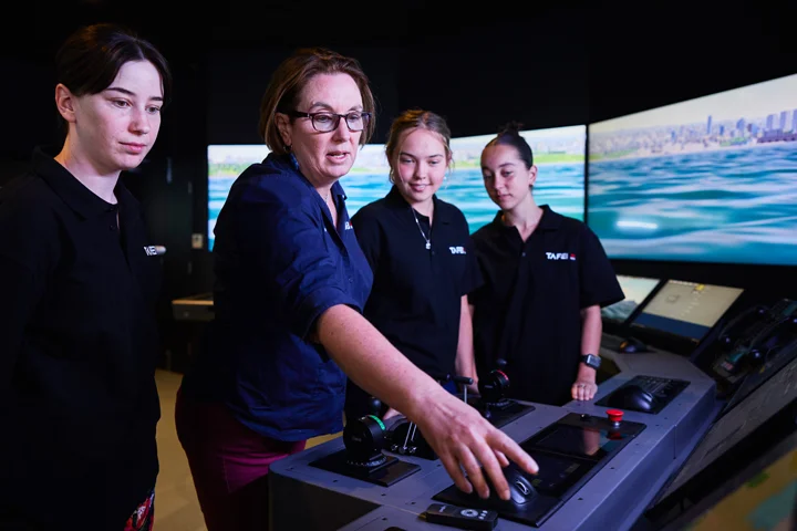 TAFE NSW maritime teacher demonstrates navigation controls to students inside the Newcastle maritime simulator, with curved floor‑to‑ceiling screens displaying an ocean and coastline scene and ship control panels positioned in front of the group for hands‑on maritime training.