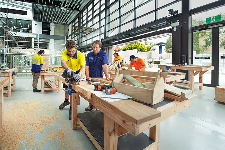 A male student, guided by his teacher, works on a carpentry project in a workshop. Two other students work on their projects in the background.