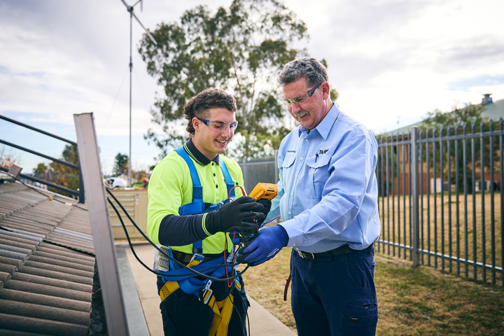 TAFE NSW renewable energy teacher guides a student wearing safety harness and high‑visibility clothing while using a digital testing device beside rooftop solar panels, demonstrating hands‑on training in safe solar installation and renewable energy systems outdoors.
