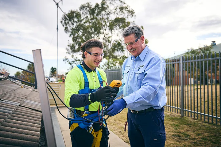 TAFE NSW renewable energy teacher guides a student wearing safety harness and high‑visibility clothing while using a digital testing device beside rooftop solar panels, demonstrating hands‑on training in safe solar installation and renewable energy systems outdoors.