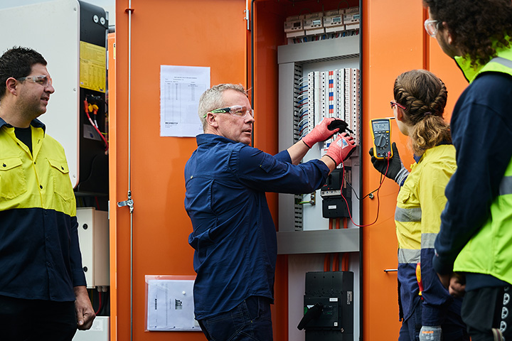 TAFE NSW electrotechnology teacher demonstrates testing procedures at an open electrical switchboard using a multimeter while students wearing safety glasses and high‑visibility clothing observe and follow instructions during hands‑on training.