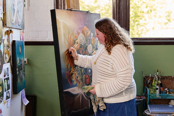 Fine arts student paints a detailed artwork on a large canvas mounted on an easel inside a studio, holding a brush and palette cloth, with other artworks, reference images, and painting tools displayed around the room and sunlight coming through tall windows.