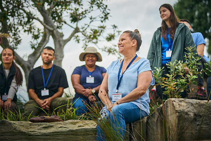 Tourism cultural guiding students sit together outdoors on rocks and grass while listening to a guide, wearing name badges in a natural bush setting with trees and native plants during a hands‑on cultural learning session.