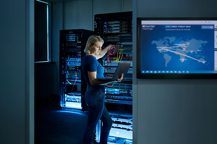 ICT student works in a cyber security server room, holding a laptop while configuring network cables inside a server rack, with illuminated equipment cabinets and a large screen displaying a live global cyber threat map in the background as part of advanced ICT training.