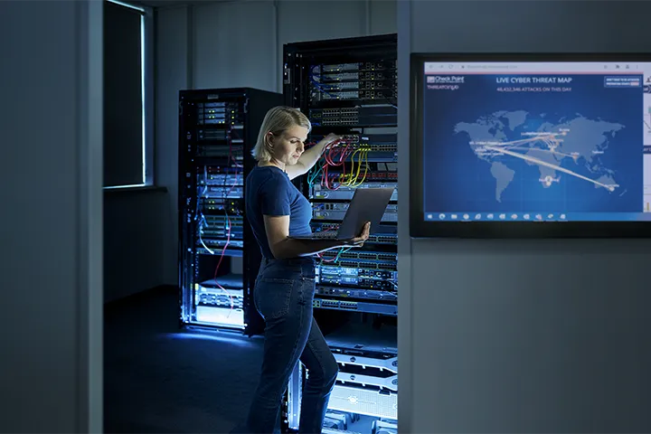 ICT student works in a cyber security server room, holding a laptop while configuring network cables inside a server rack, with illuminated equipment cabinets and a large screen displaying a live global cyber threat map in the background as part of advanced ICT training.