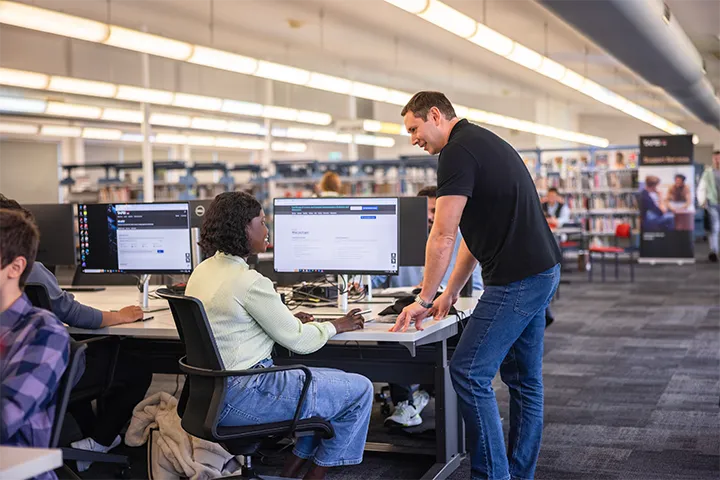 A man is helping a woman at the library. She is seated at a desktop computer and he is standing next to her. They are smiling and engaged in conversation. There are other students working in the background.