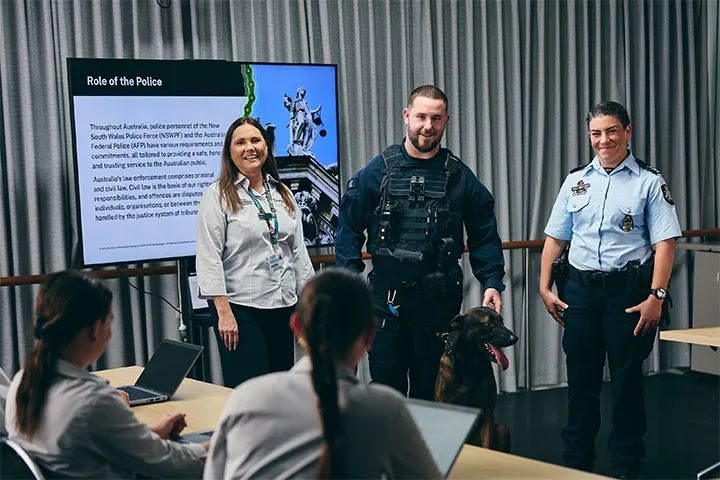TAFE NSW IPROWD classroom session where two NSW Police Officers, one holding a dog, and a woman are presenting to students seated at desks with laptops. A screen behind them displays the title 'Role of the Police' along with information about law enforcement responsibilities in Australia.