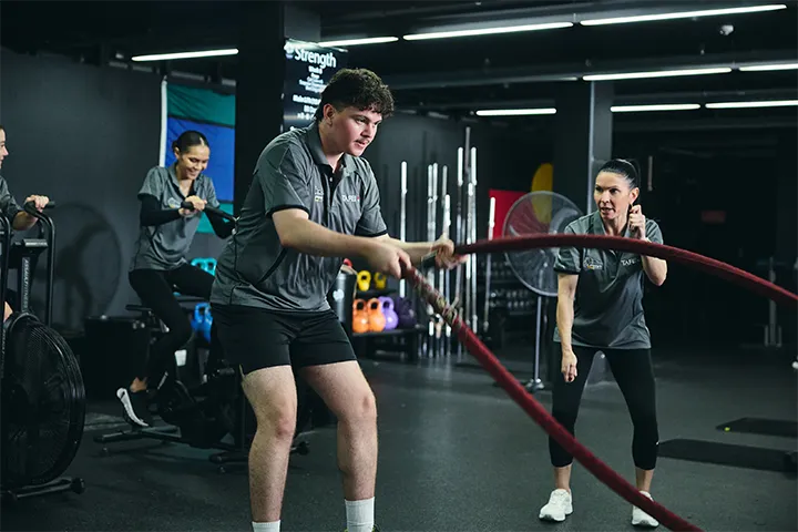 Participants in IPROWD training at a gym, with one person using battle ropes, another providing instruction, and others on exercise bikes, surrounded by kettlebells, weight racks, and a 'Strength' sign in the background.