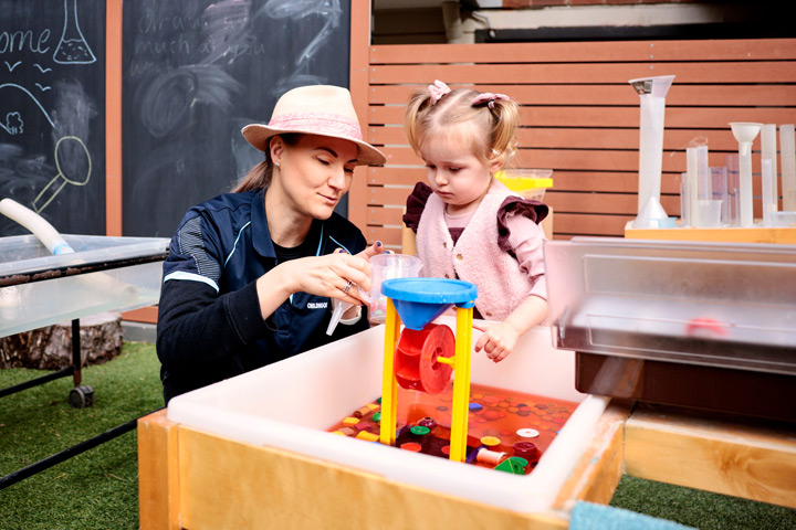 TAFE NSW Early Childhood Education and Care student supports a young child during hands‑on play‑based learning at a water sensory table, guiding the use of measuring cups and colourful funnels while educational play equipment and chalkboard activities are visible in an outdoor learning area.