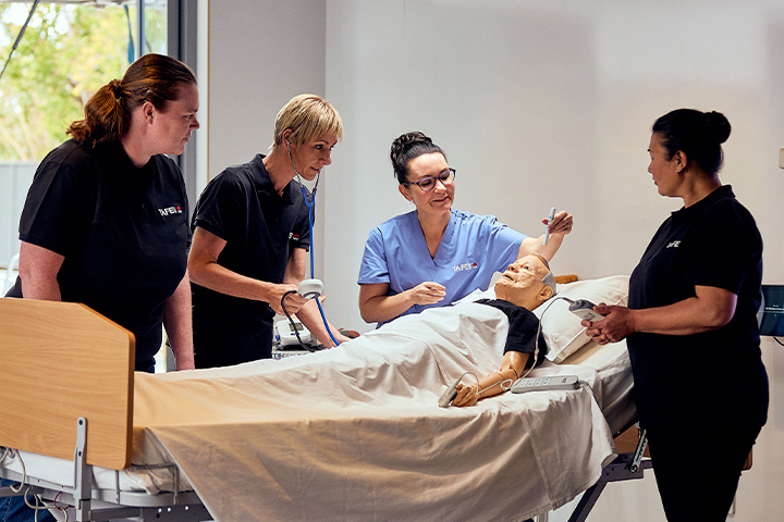 Three students and a teacher stand around a mannequin in a hospital bed in a training facility. Two of the students and the teacher are holding different pieces of medical equipment. Everyone is watching the teacher hold an item over the mannequin’s head.   