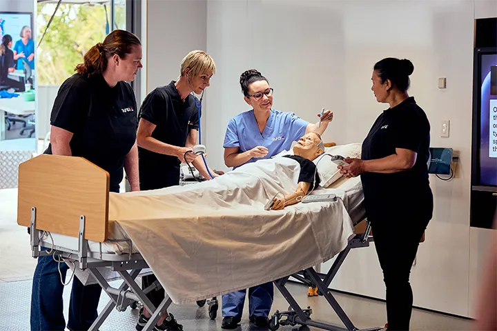 Three students and a teacher stand around a mannequin in a hospital bed in a training facility. Two of the students and the teacher are holding different pieces of medical equipment. Everyone is watching the teacher hold an item over the mannequin’s head.   