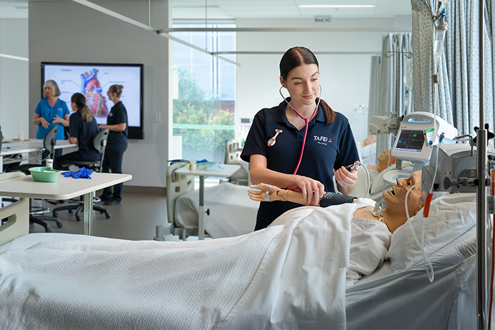 A nursing student practices on a dummy in a medical bed. In the background, a teacher presents to other nursing students.