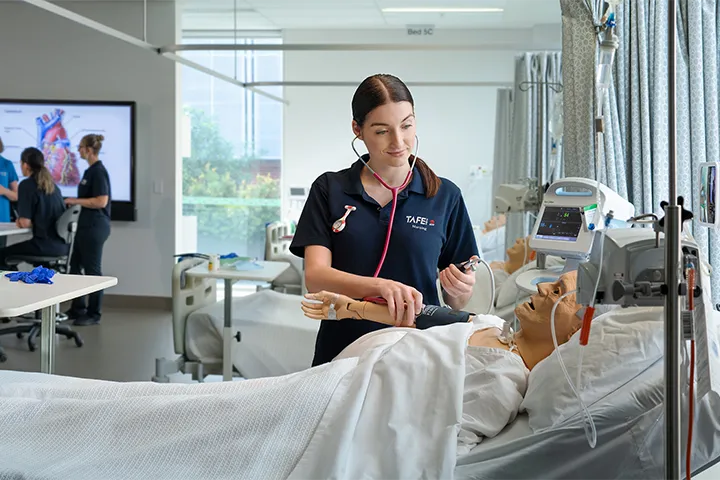 A nursing student practices on a dummy in a medical bed. In the background, a teacher presents to other nursing students.