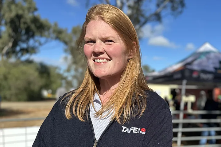 TAFE NSW Head Teacher of Agriculture and Animal Care, Anna Ingold, is standing outside in the sun, smiling at the camera. She wears a navy-blue TAFE NSW jacket. In the background there is blue sky, trees and a marquee.