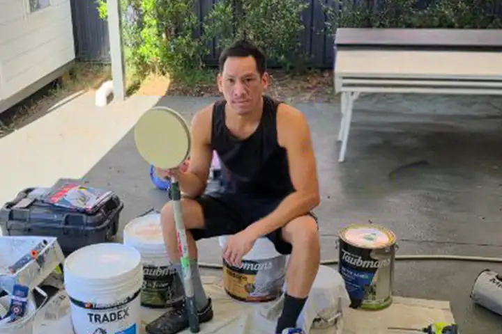 Anthony Phonlamuang sits on a paint bucket in an undercover outdoor area with a painting tool in his hand. He’s wearing a black singlet and shorts splattered with white paint. He is surrounded by buckets of paint and painting equipment.
