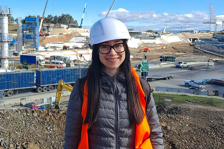 Aza Rondon stands on a construction site in a winter jacket, and white hard hat, smiling at the camera. 
