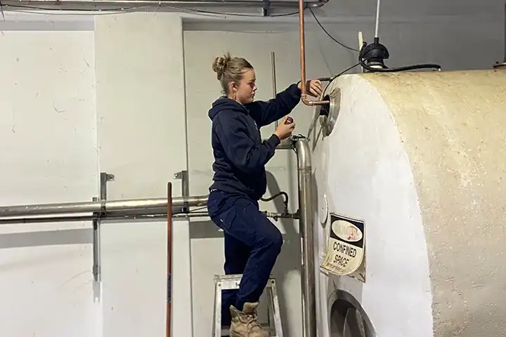Callie Hagan, a female apprentice stands on a ladder working on a pipe connection at the top of a fibreglass storage tank. She wears work boots, and navy-blue trousers and a hoodie. 