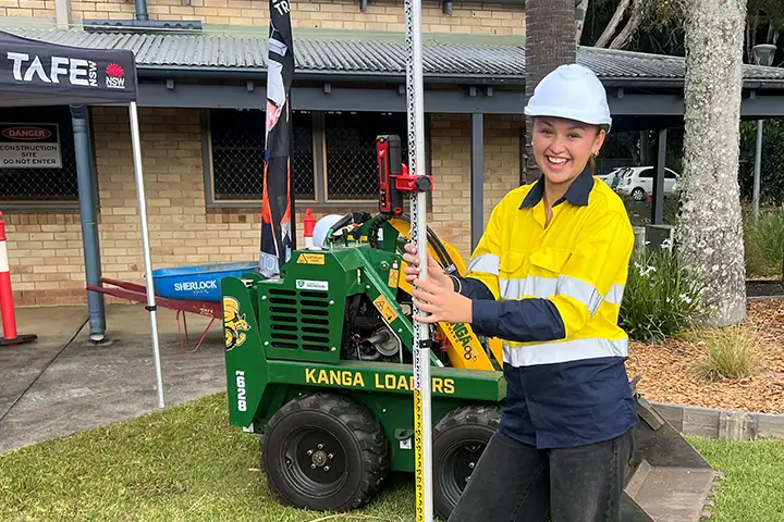 17-year-old Charlotte Hape stands in the grounds of TAFE NSW Ballina, holding a measuring tool. She wears a yellow high-vis shirt, a white hard hat and is smiling at the camera.