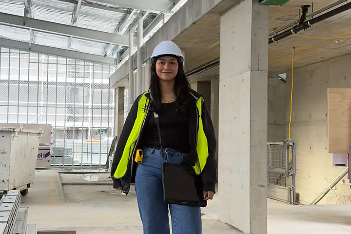 Andrea Ramiez Pertuz stands in the centre of an indoor construction site. She’s wearing blue jeans, a black shirt, yellow high-vis vest and white hard hat. She has long black hair and is smiling at the camera.