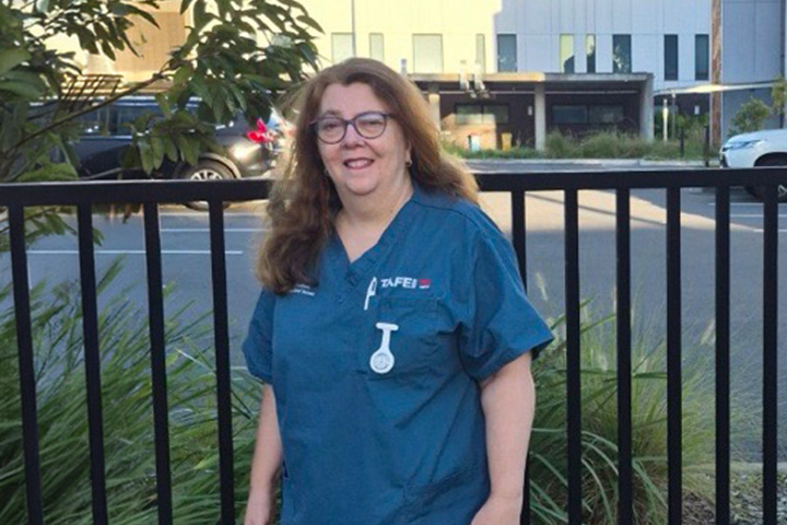 Denise Beckman stands on a patch of lawn in front of a black metal fence. She wears a TAFE NSW nursing uniform and smiles at the camera. The Sutherland Hospital is in the distance against a backdrop of blue sky.