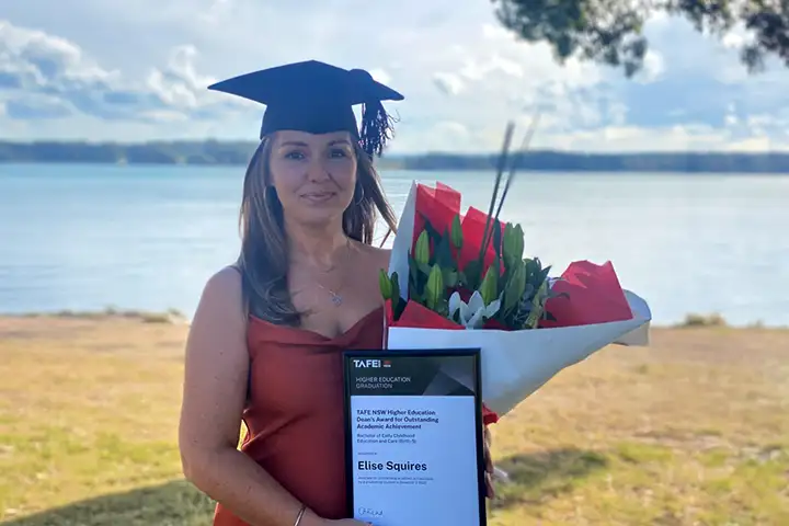 TAFE NSW graduate Elise Squires smiles at the camera, wearing a black graduation cap with tassel and a red dress. She holds a bunch of flowers and her award for outstanding academic achievement. She’s standing outside in the shade of a tree, and there is a river in the background.