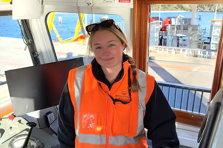 Photo of Ella Field smiling on a moored boat’s command deck in an orange high-vis jacket on a sunny day, with calm blue water behind her.