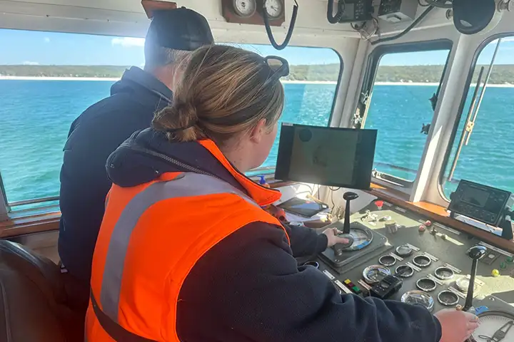 Ella Field stands beside a male colleague at a boat’s command deck. Both have their back to the camera. She wears an orange high-vis jacket with her hair tied in a knot. He wears a dark jacket and a cap and stands in front of a monitor. It’s a sunny day and parkland and beaches are in the distance.
