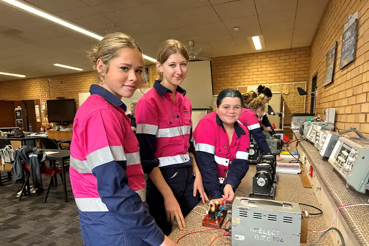 Zanthia Fulton, Amaya Rutherford and Lilly James are standing in a TAFE NSW classroom, gathered around some electrical equipment on a bench. They are wearing pink and navy blue high-vis shirts, and are looking towards the camera, smiling. 